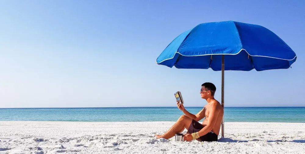 Man reading a paperback book under a beach umbrella.